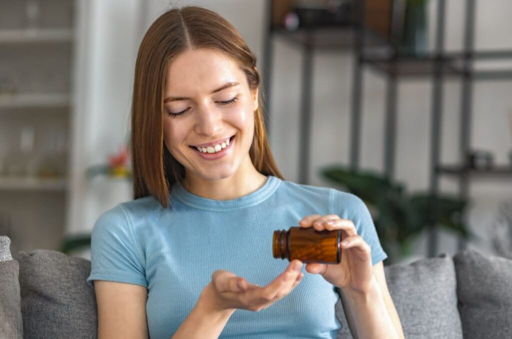 A young person sitting on a couch and smiling while holding a brown bottle of vitamins for eye health.