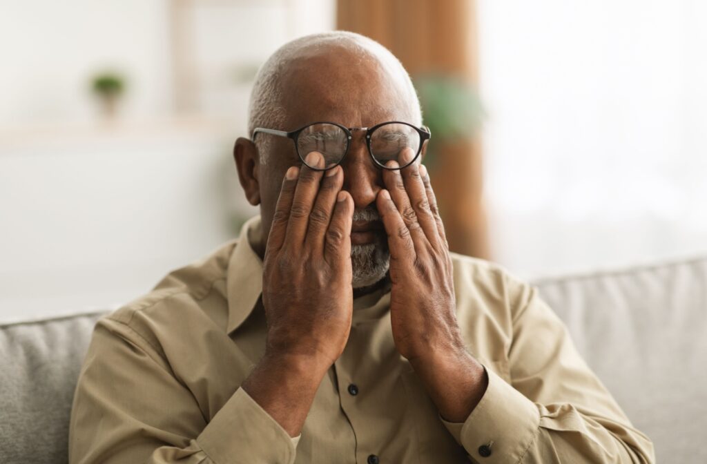 An older adult on their couch at home lifting their glasses and rubbing their eyes, dealing with discomfort and flashing lights.