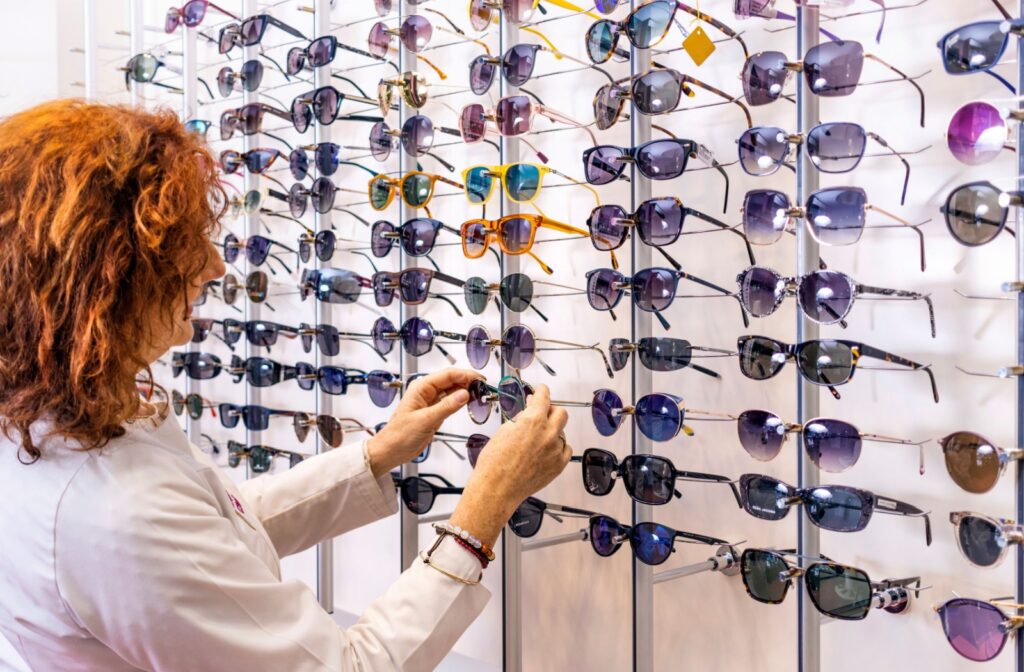 A person in a white coat selects a pair of sunglasses from a wall display filled with various styles.