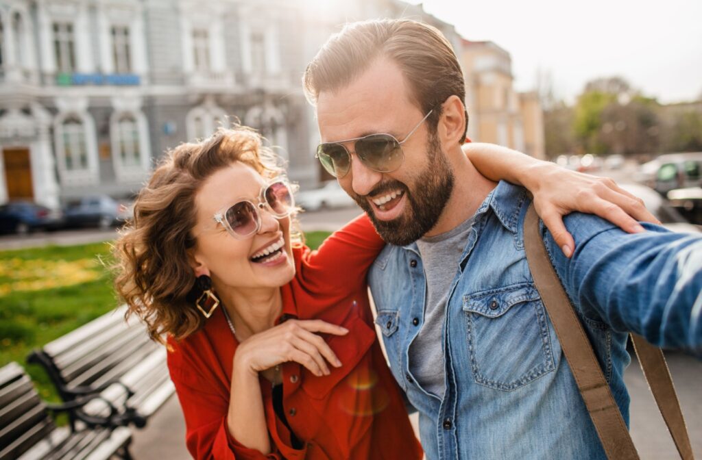 A smiling couple wearing sunglasses walks together on a sunny day in the city.