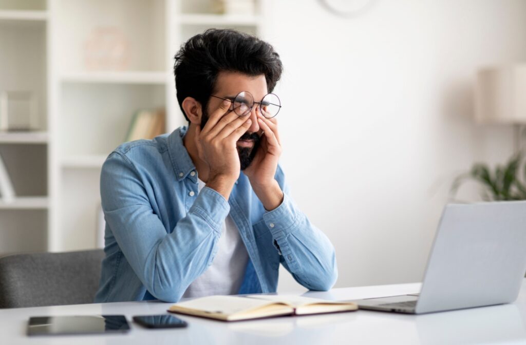 A person sitting a desk in front of a laptop screen, rubbing their eyes due to eye strain.