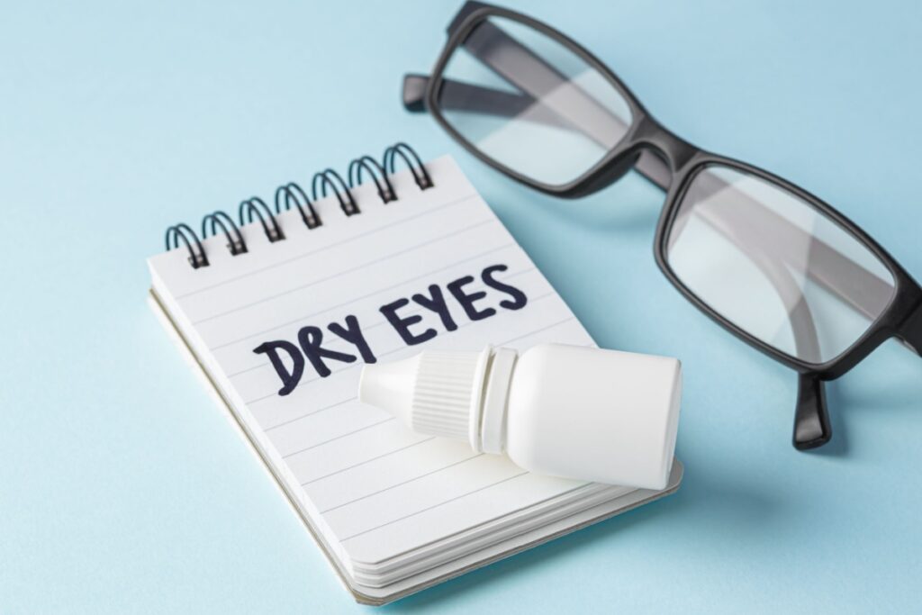 A notepad with the words "Dry Eyes" on it on a blue background with glasses and eye drops beside.